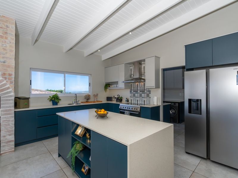 Modern kitchen featuring a central island, stone countertops, built-in appliances, exposed beam ceiling, and an integrated brick fireplace, with an adjacent scullery area.