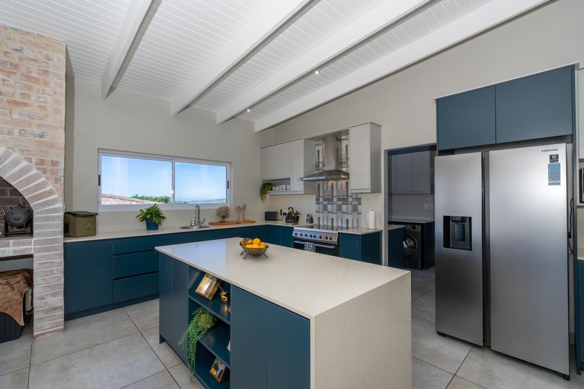 Modern kitchen featuring a central island, stone countertops, built-in appliances, exposed beam ceiling, and an integrated brick fireplace, with an adjacent scullery area.