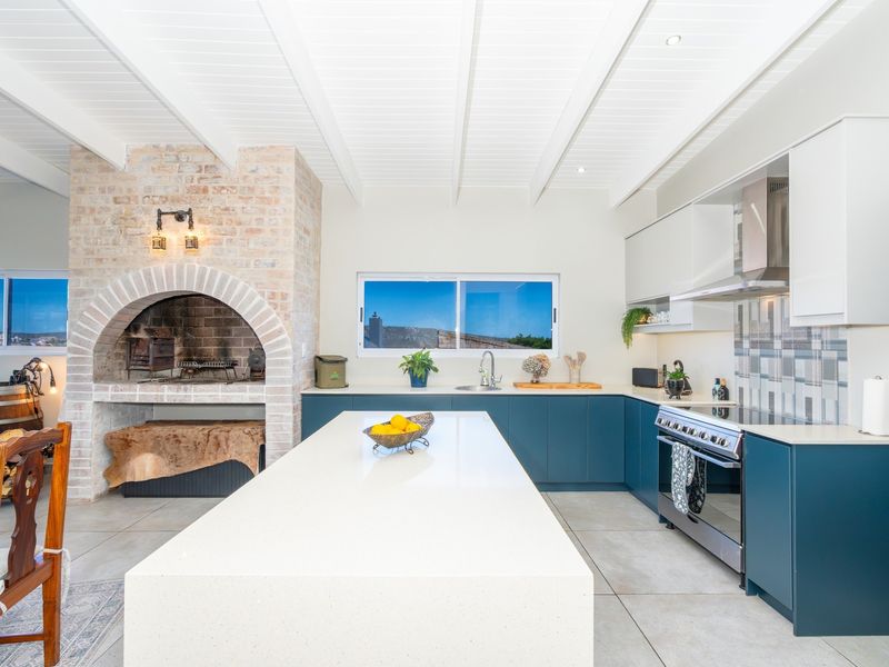 Modern open-plan kitchen featuring a large central island, quartz countertops, built-in oven, extractor fan, and a distinctive brick fireplace, all under a bright beamed ceiling.