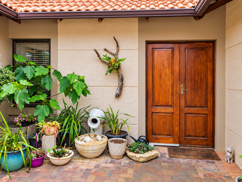A well-maintained covered patio area featuring brick paving, a double wooden entrance door, and terracotta roof tiling.