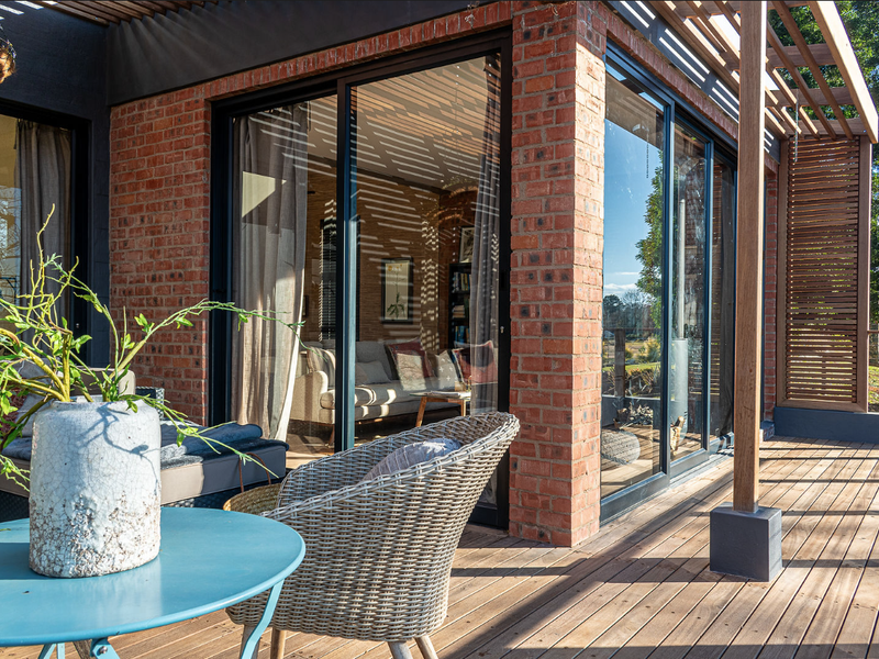 A modern covered patio featuring timber decking, exposed brick walls, and a timber pergola, seamlessly connecting to the interior living space through sliding glass doors.
