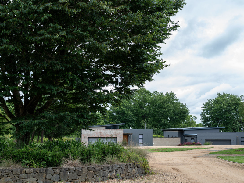 A view of a modern residential property featuring a stone retaining wall, mature landscaping, and a gravel driveway leading to the garage.
