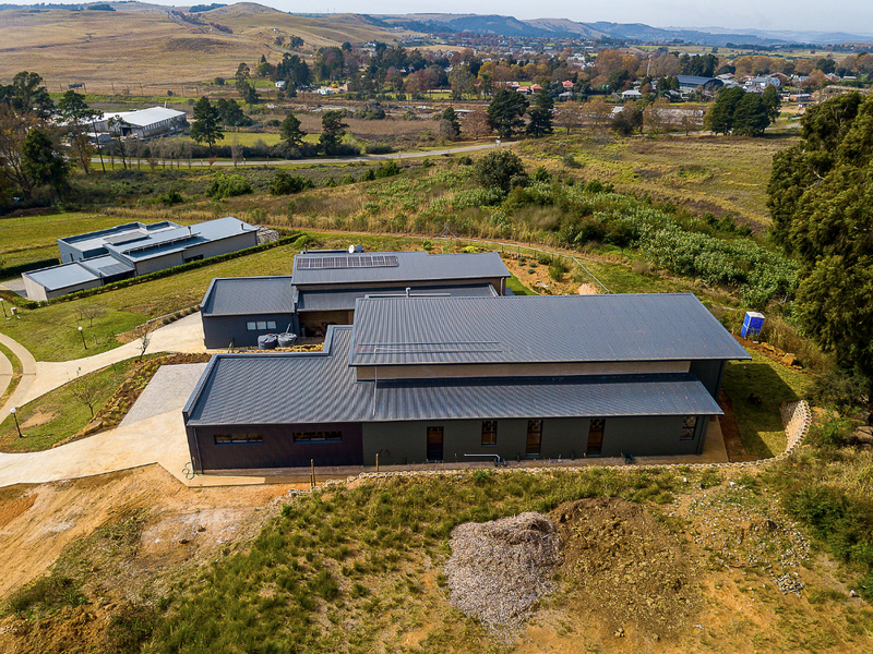 Aerial view of a modern house with a corrugated metal roof, solar panels, and surrounding rural landscape.