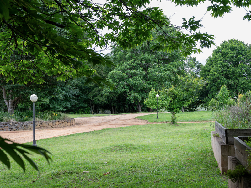 A well-maintained garden area featuring a gravel driveway, stone retaining wall, timber sleeper garden beds, and established mature trees.