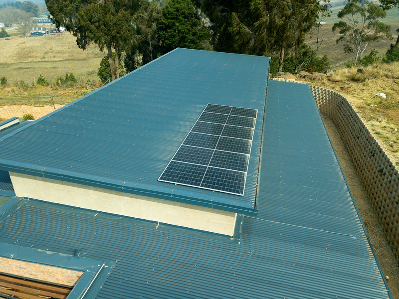 Aerial view of a modern residence featuring a corrugated metal roof with integrated solar panels and a structured retaining wall.