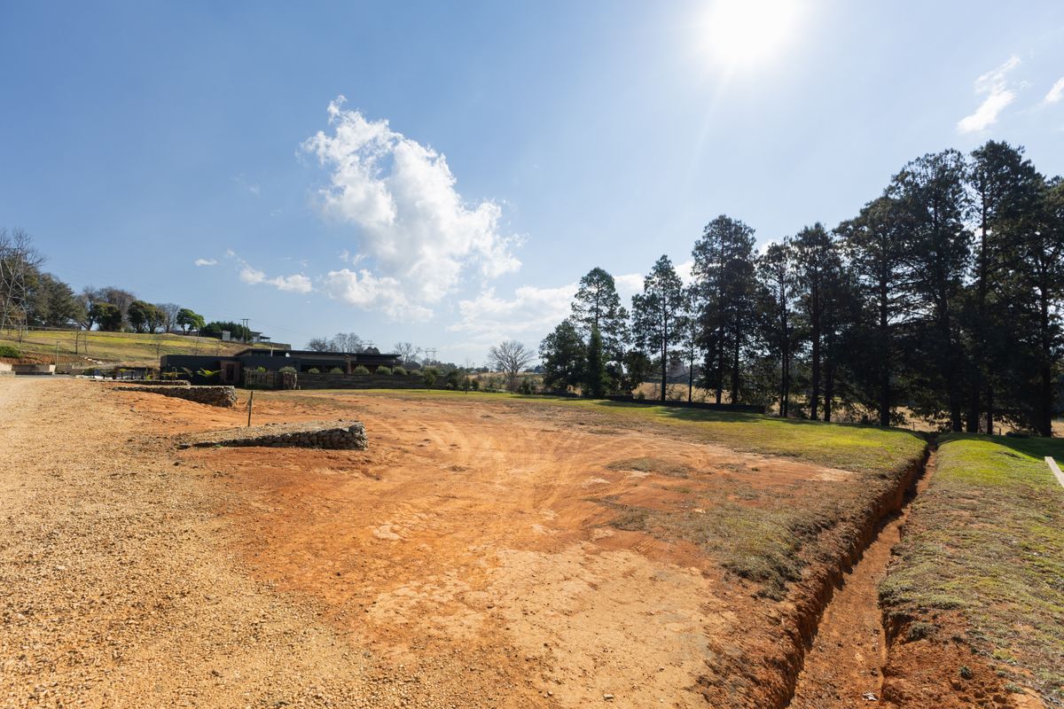 A view of a vacant residential plot featuring cleared earth, a small stone retaining wall, and mature trees along the perimeter.