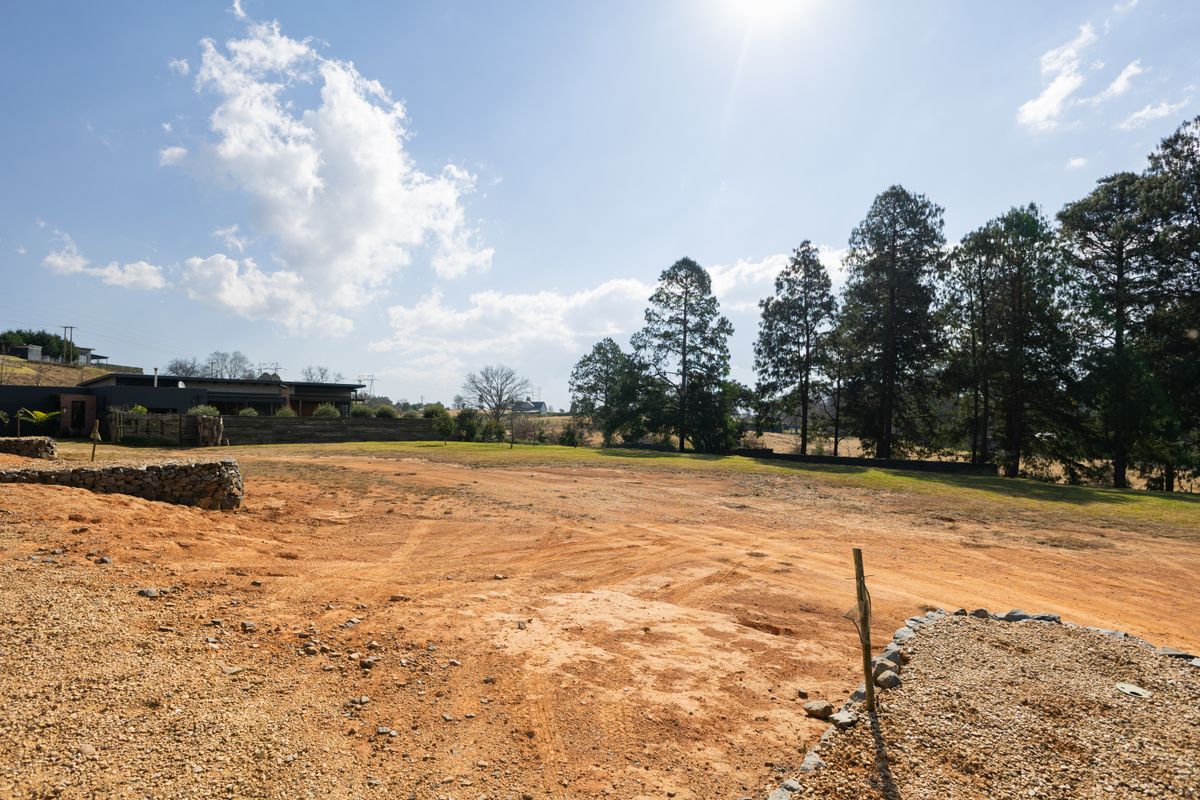A view of a vacant residential plot featuring a gravel area, retaining wall, and mature trees in the background.