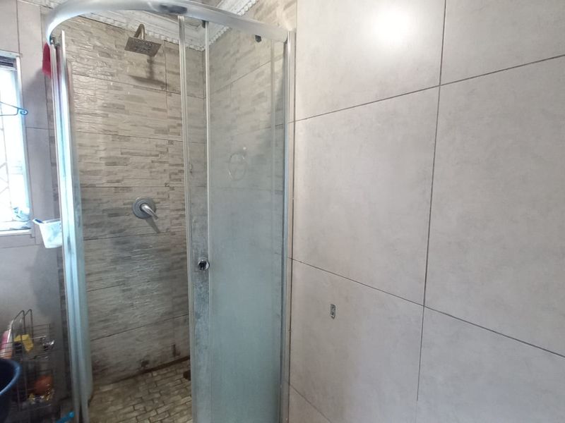 A functional bathroom featuring a corner shower enclosure with tiled walls, a pedestal basin, and a window with security bars.