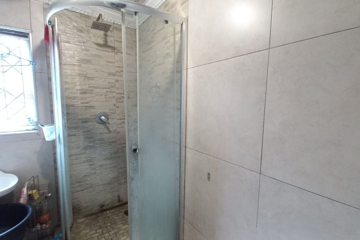 A functional bathroom featuring a corner shower enclosure with tiled walls, a pedestal basin, and a window with security bars.