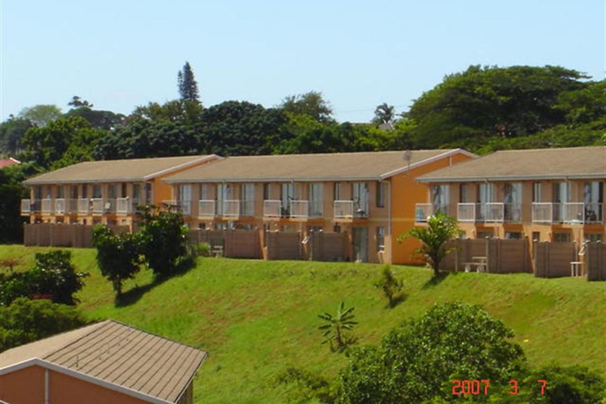 A view of the rear elevation of a multi-unit residential complex featuring balconies, sliding doors, and private fenced courtyards set against a landscaped hillside.