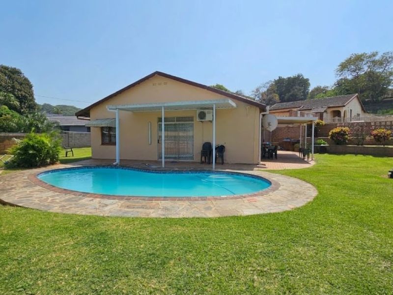 Rear view of the property featuring a swimming pool with stone coping, a covered patio area, and a well-maintained garden.