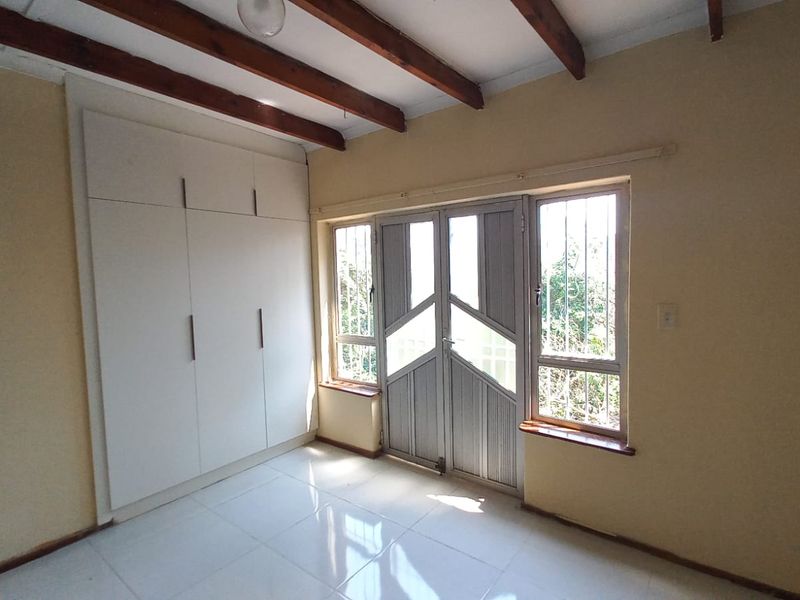 A bright, empty bedroom featuring built-in cupboards, tiled flooring, and exposed timber ceiling beams, with direct access to the exterior via a security gate.