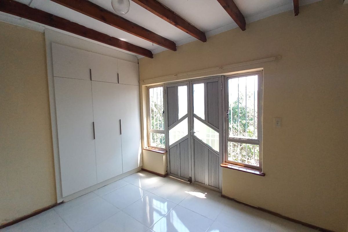 A bright, empty bedroom featuring built-in cupboards, tiled flooring, and exposed timber ceiling beams, with direct access to the exterior via a security gate.