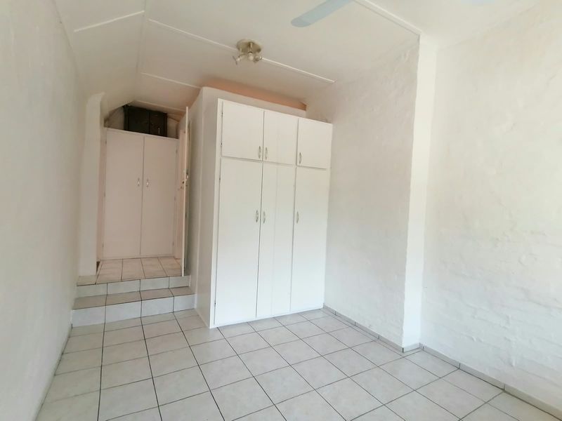 A simple bedroom featuring white built-in cupboards, tiled flooring, and a ceiling fan, offering a clean and functional space.
