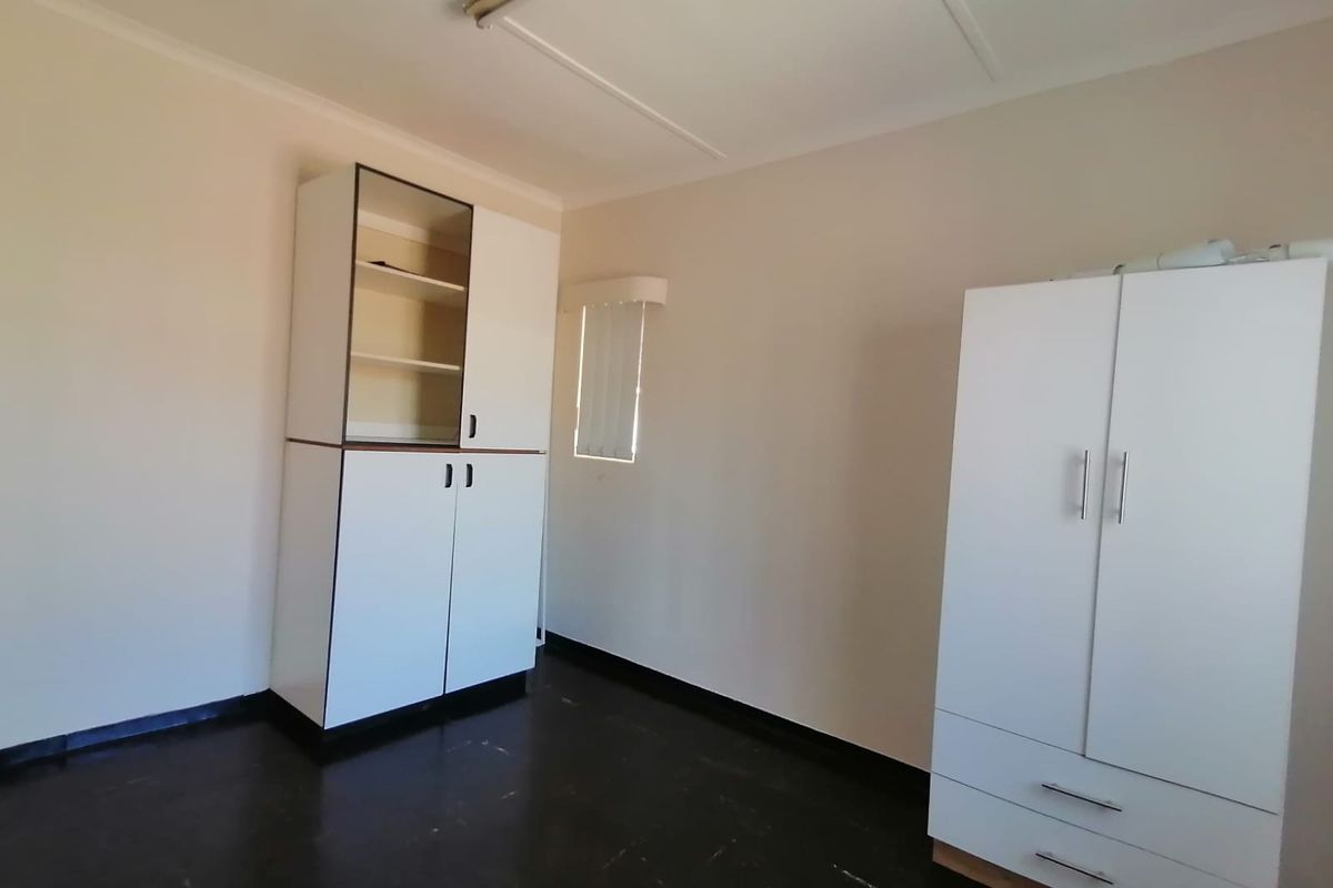 A clean, empty bedroom featuring built-in storage cupboards, dark vinyl flooring, and a window with vertical blinds.