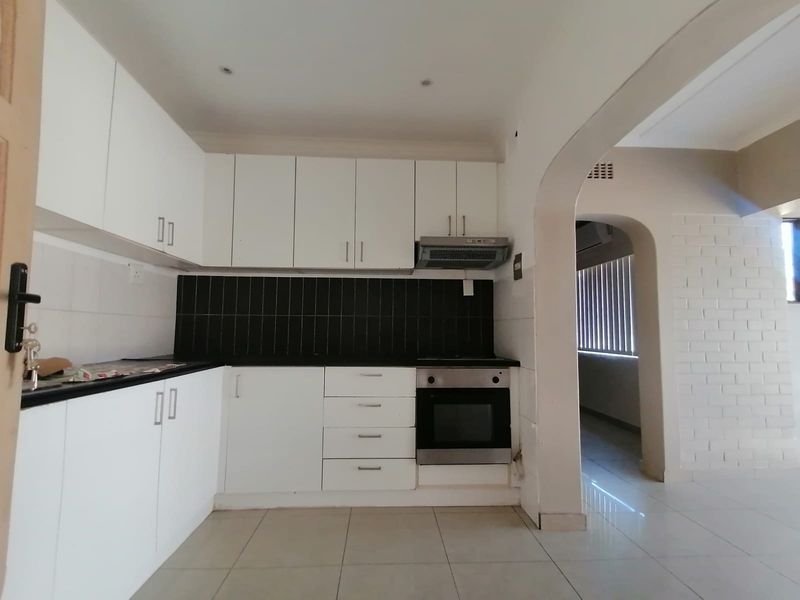 A clean, modern kitchen featuring white cabinetry, a black tiled backsplash, built-in oven, and extractor fan, leading into an adjacent living area through an archway.