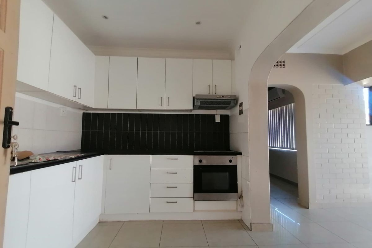 A clean, modern kitchen featuring white cabinetry, a black tiled backsplash, built-in oven, and extractor fan, leading into an adjacent living area through an archway.