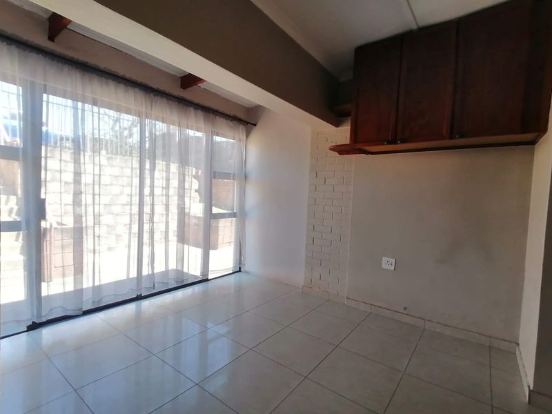 An empty living area featuring tiled floors, exposed ceiling beams, a brick feature wall, and large sliding glass doors leading to the exterior.