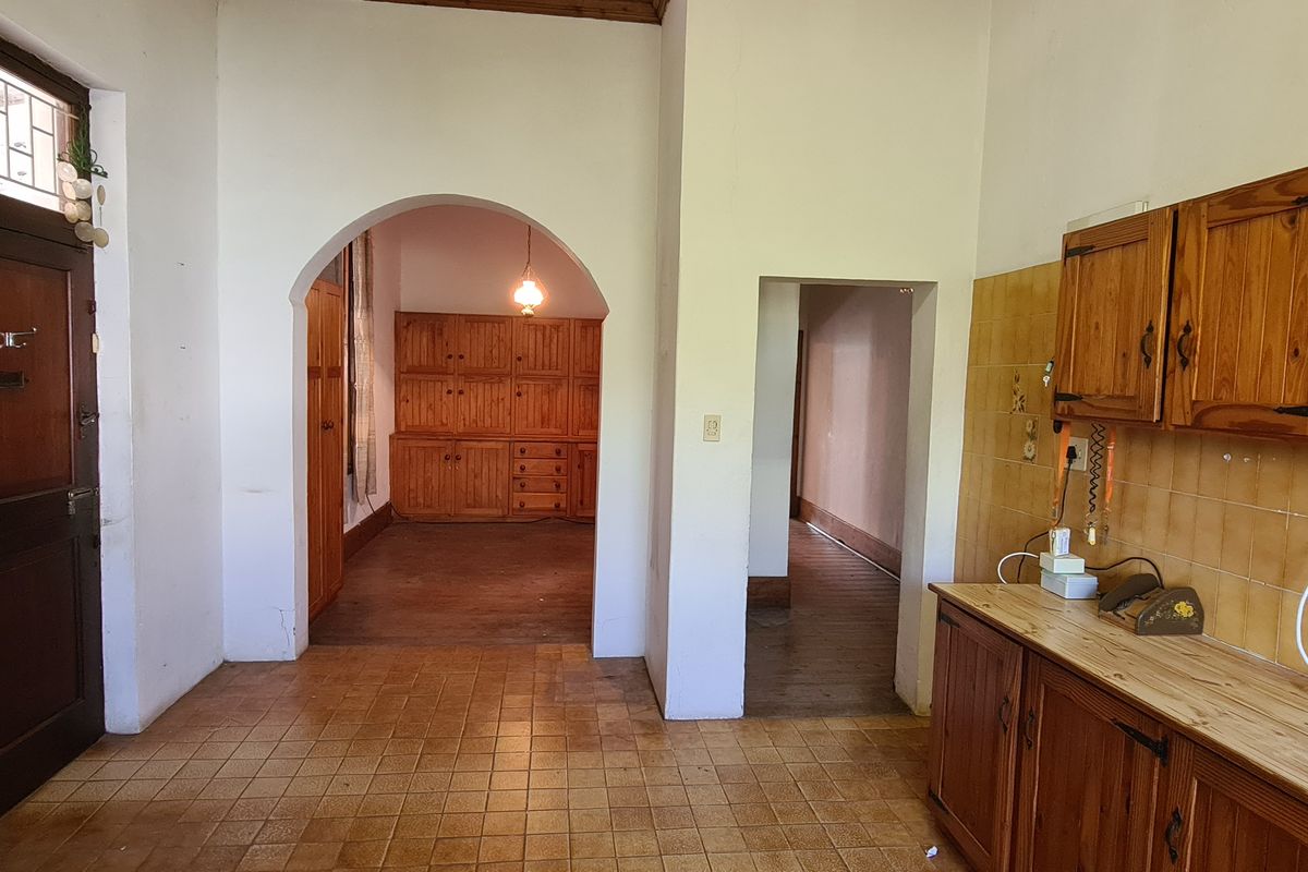 A dated kitchen featuring wooden cabinetry, tiled splashbacks, and tiled flooring, with an arched doorway leading to an adjacent living area.