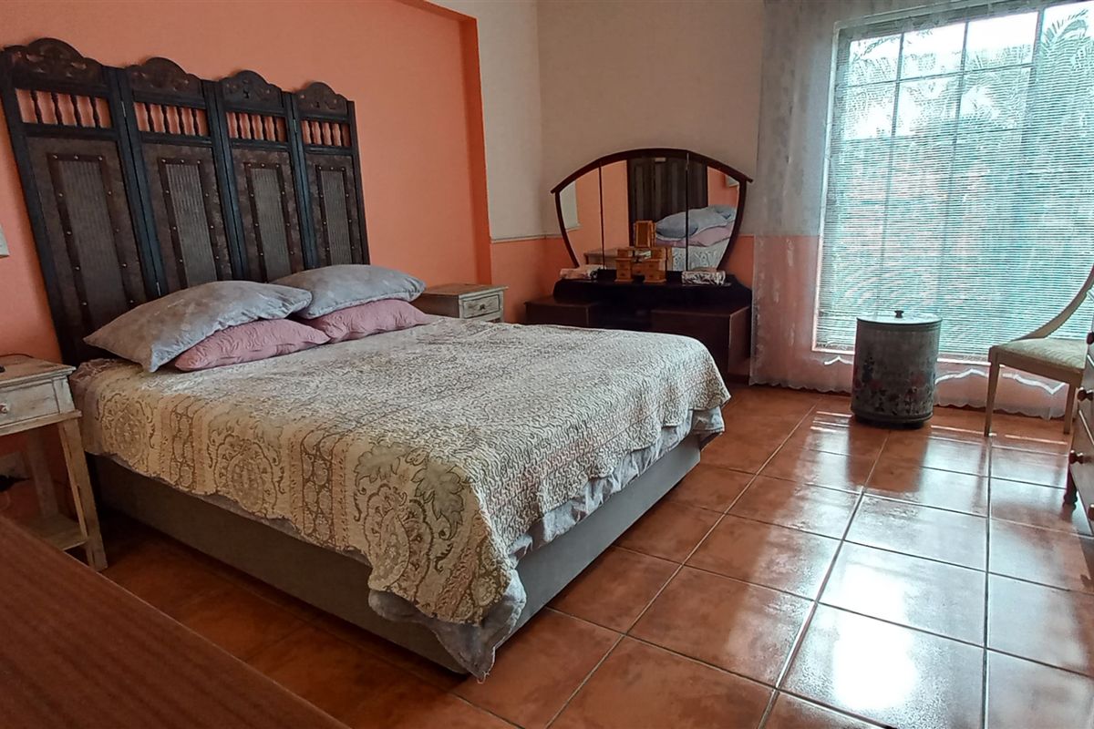 A spacious bedroom featuring terracotta floor tiles, a large window with natural light, and a prominent decorative headboard.