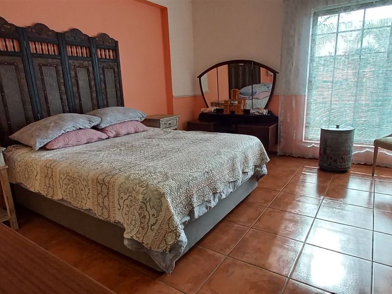 A spacious bedroom featuring terracotta floor tiles, a large window with natural light, and a prominent decorative headboard.