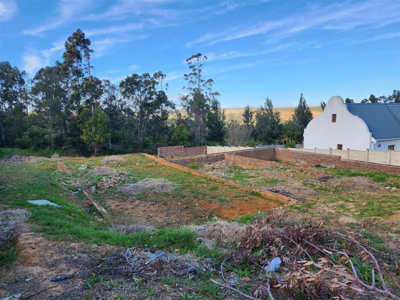 A vacant residential plot featuring existing brick foundation walls and a boundary wall, set against a backdrop of mature trees and neighboring property.