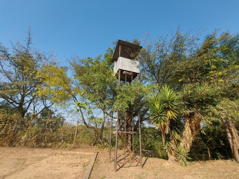 An elevated metal observation tower situated within a garden area, featuring a covered platform and surrounding mature trees.