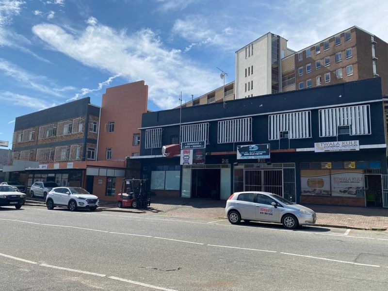 Street-level view of a multi-story commercial building featuring various storefronts, security shutters, and paved sidewalk access in an urban setting.