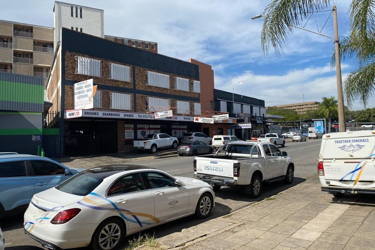 Street-level view of a commercial building featuring multiple storefronts with security shutters and signage, situated along a paved sidewalk.