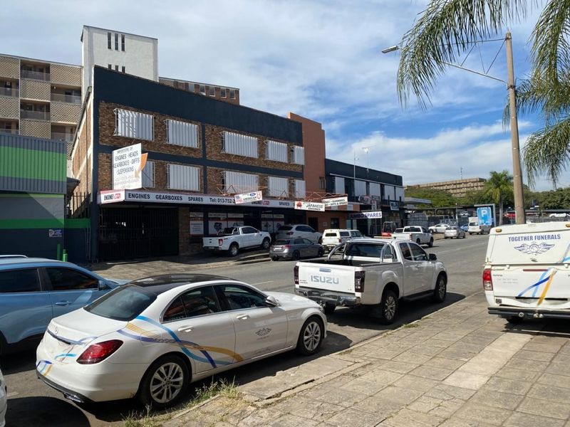 Street-level view of a commercial building featuring multiple storefronts with security shutters and signage, situated along a paved sidewalk.