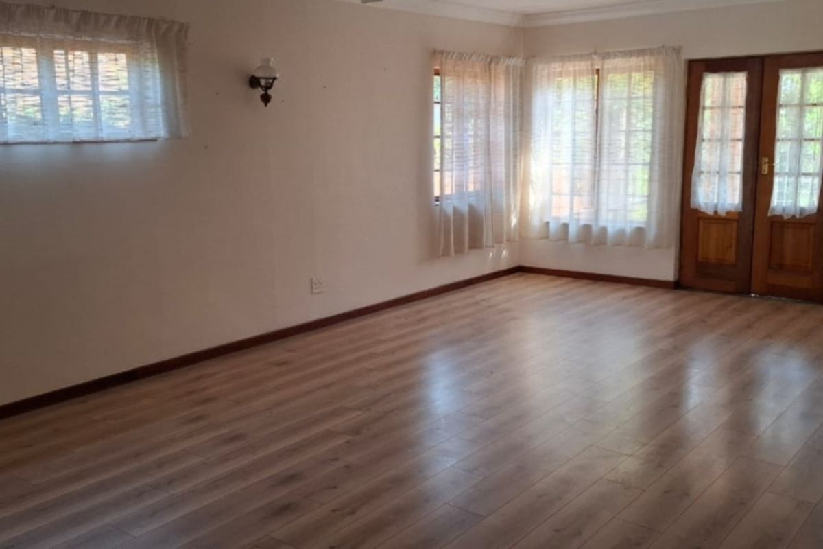 A spacious, empty living room featuring light-toned laminate flooring, neutral walls, and wooden double doors leading to the exterior.