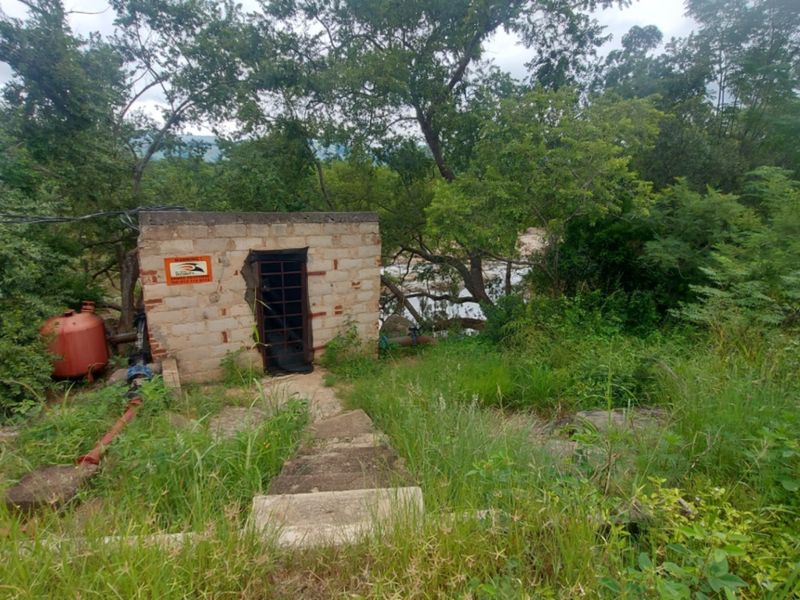 A small, utilitarian concrete block structure situated in a heavily overgrown area, featuring a metal security gate and external plumbing infrastructure.