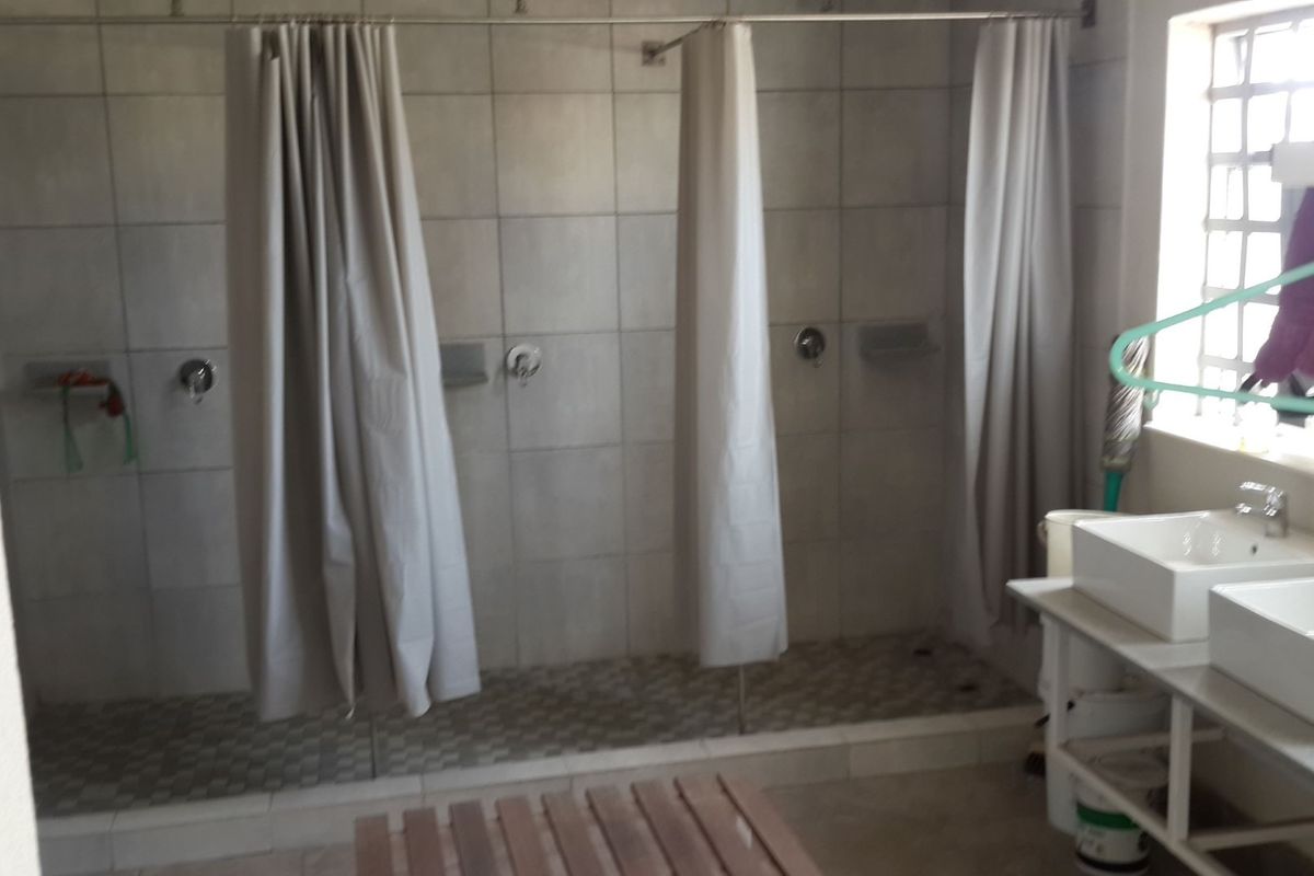 Industrial-style communal bathroom featuring multiple shower stalls with tiled flooring and a dual vessel sink vanity unit.