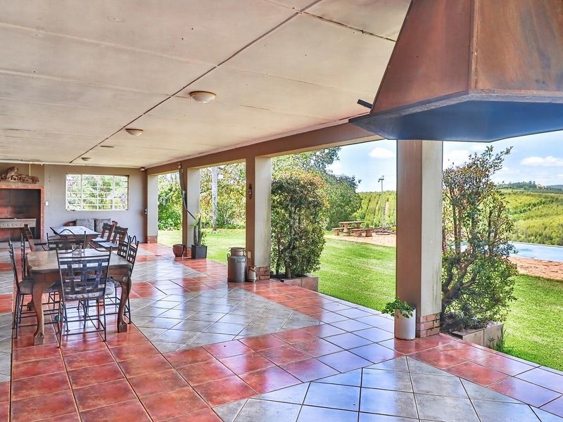Spacious covered patio featuring a built-in braai, suspended fireplace hood, and tiled flooring, overlooking a lush garden and swimming pool area.