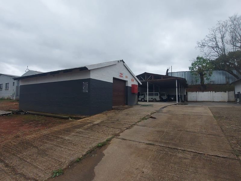 Industrial property exterior featuring a spray booth unit, concrete driveway, and covered parking area with weathered brick and metal structures.