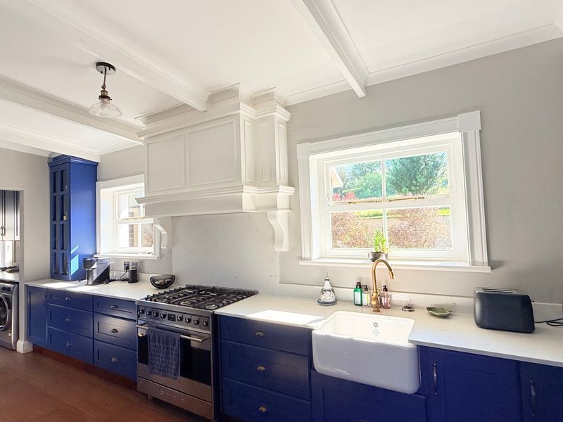 A well-appointed kitchen featuring blue cabinetry, stone countertops, a farmhouse sink, and a gas range with a custom extractor hood.