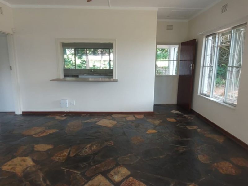 An empty living room featuring rustic stone flooring, a convenient serving hatch to the kitchen, and large windows with security bars.