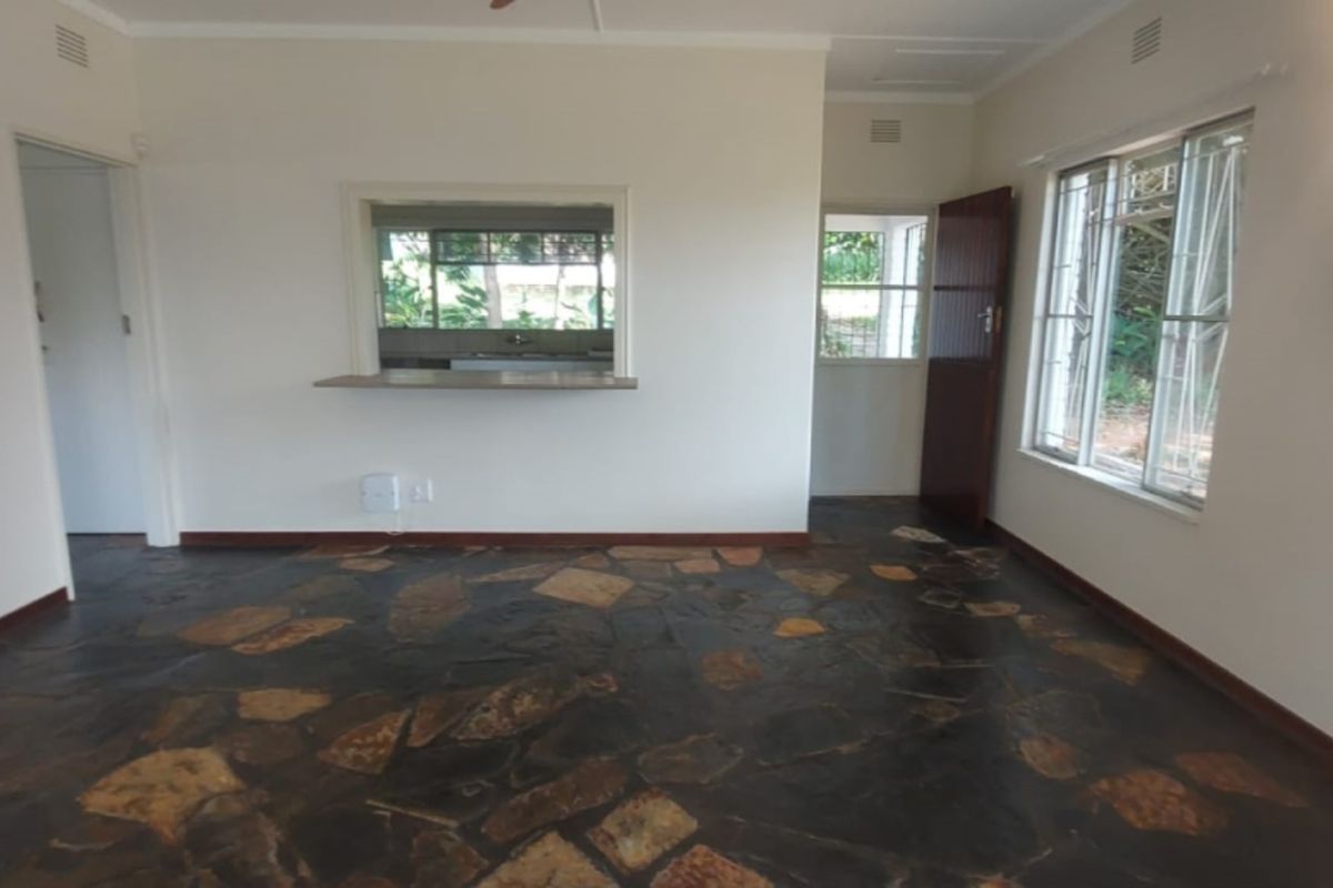 An empty living room featuring rustic stone flooring, a convenient serving hatch to the kitchen, and large windows with security bars.