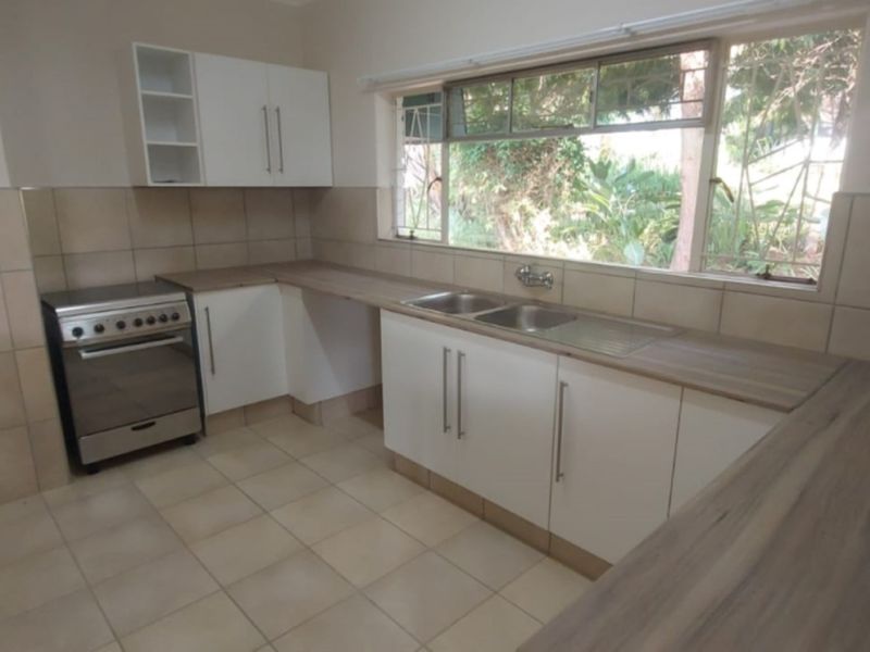 A functional kitchen featuring white cabinetry, laminate countertops, a stainless steel double sink, and a freestanding stove, with tiled finishes throughout.