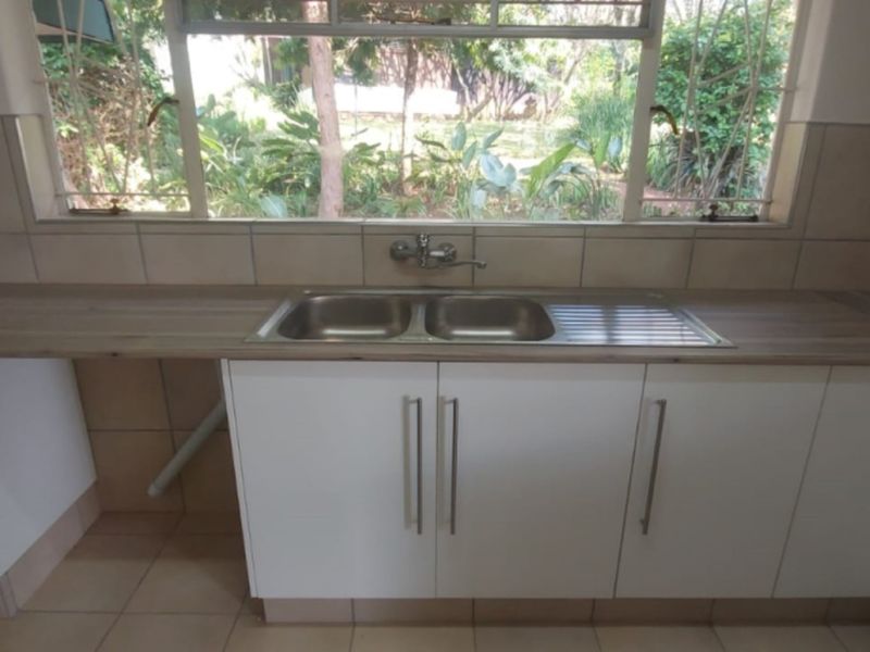 A functional kitchen area featuring a double stainless steel sink, laminate countertops, tiled backsplash, and white cabinetry with a view of the garden.