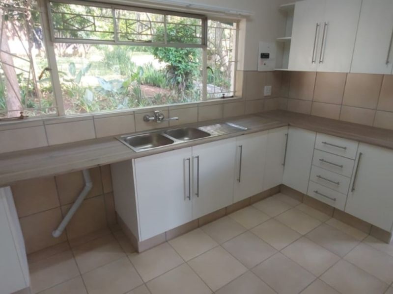 A clean, functional kitchen featuring white cabinetry, laminate countertops, tiled backsplash, and a double basin sink overlooking the garden.