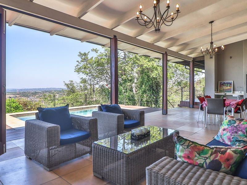 Spacious covered patio featuring a built-in braai, tiled flooring, and exposed beam ceilings, seamlessly opening to a swimming pool and scenic outdoor views.