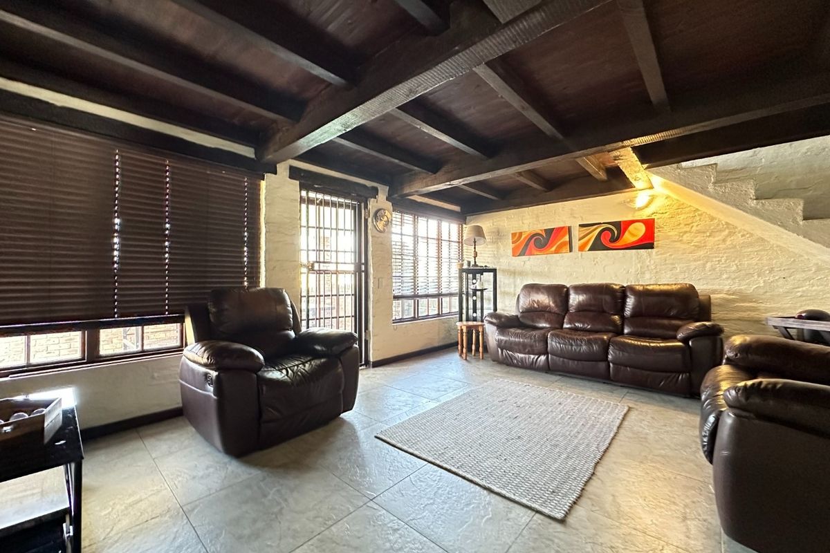A rustic living room featuring exposed dark timber ceiling beams, textured white brick walls, and tiled flooring with natural light from large windows.