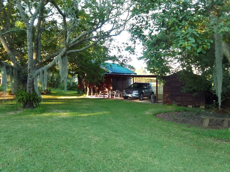 A view of the property exterior featuring a lush green garden, mature trees, and a covered parking area adjacent to the main dwelling.