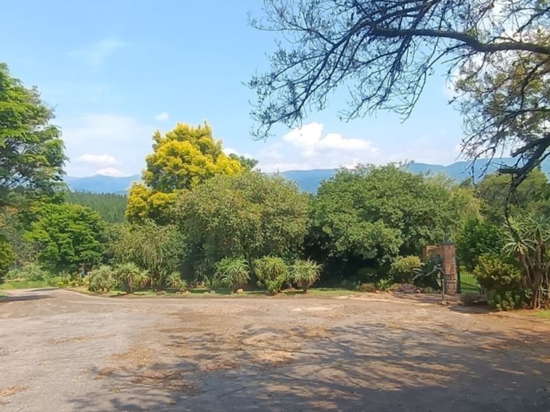 A wide view of the property entrance featuring a paved driveway, mature trees, and established garden landscaping with mountain views in the background.