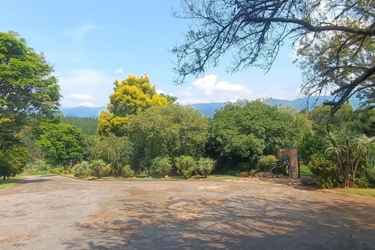 A wide view of the property entrance featuring a paved driveway, mature trees, and established garden landscaping with mountain views in the background.