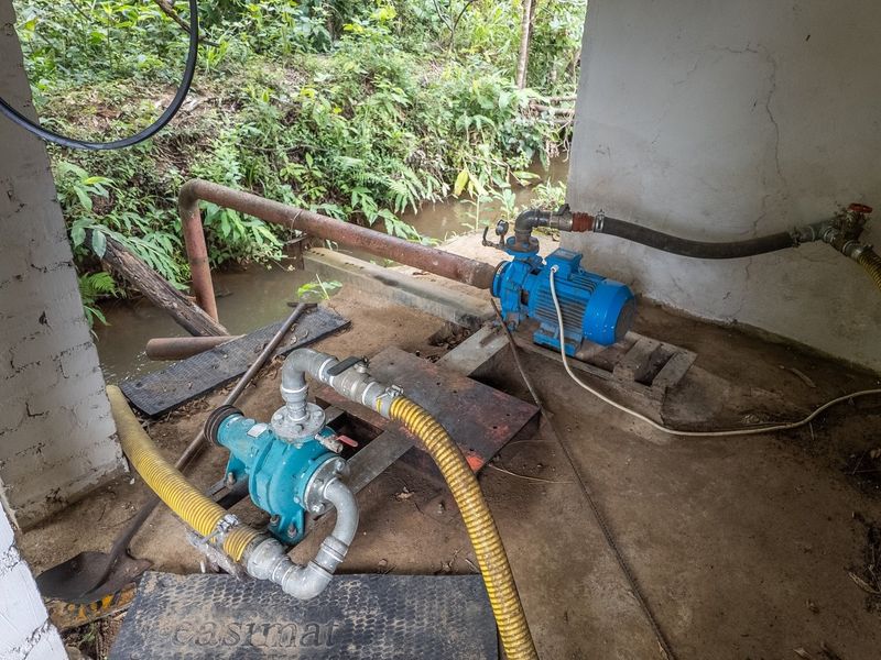 Utility area housing electric water pump infrastructure with connected plumbing and concrete flooring, situated in a semi-enclosed structure.