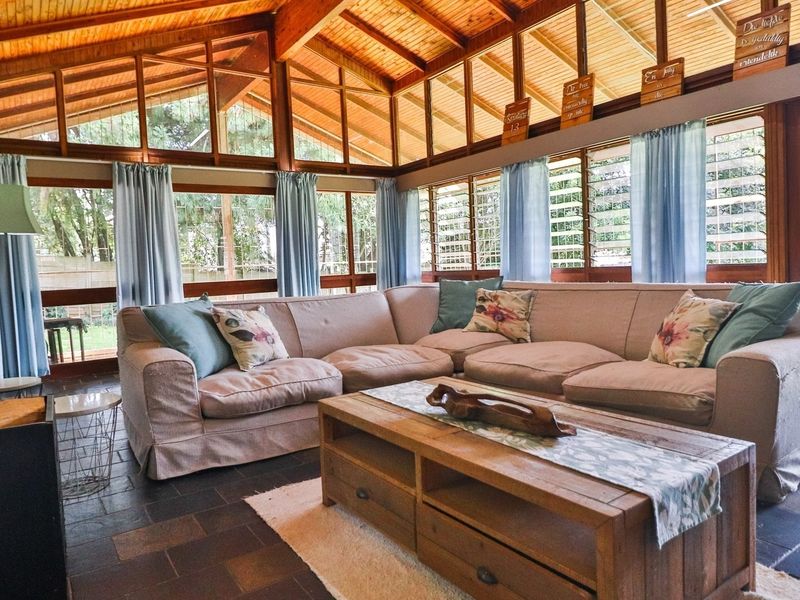 Spacious living room featuring a high exposed timber beam ceiling, stone fireplace, and large windows providing ample natural light and garden views.