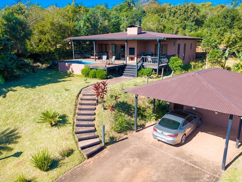 Aerial view of a residential property featuring a house with a timber deck, swimming pool, carport, and landscaped garden set in a natural environment.