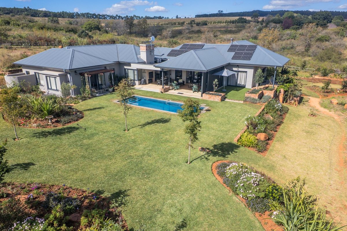 Aerial view of a modern single-story residence featuring a swimming pool, covered patio, solar panels, and extensive landscaped gardens in a rural setting.